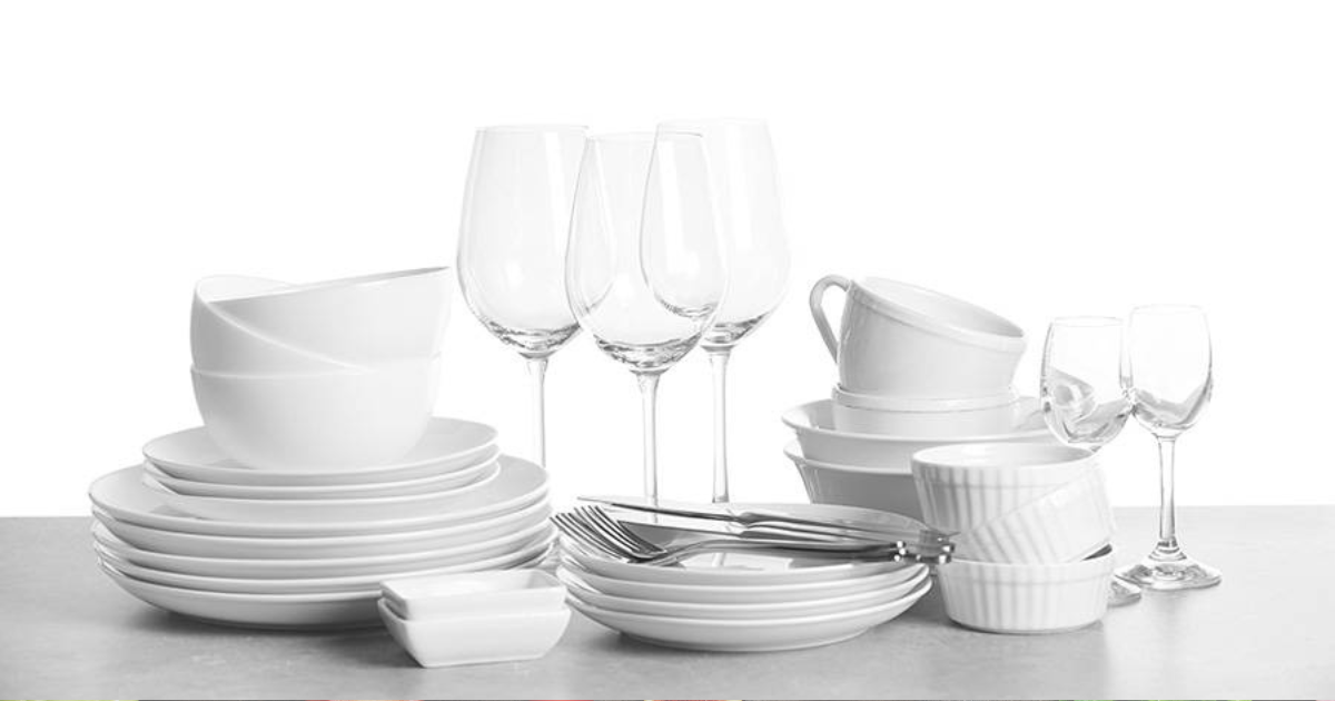 Une collection de plats, de bols, de tasses et de verres blancs propres, y compris des verres à vin et des ramequins, soigneusement empilés sur une surface grise sur un fond blanc.