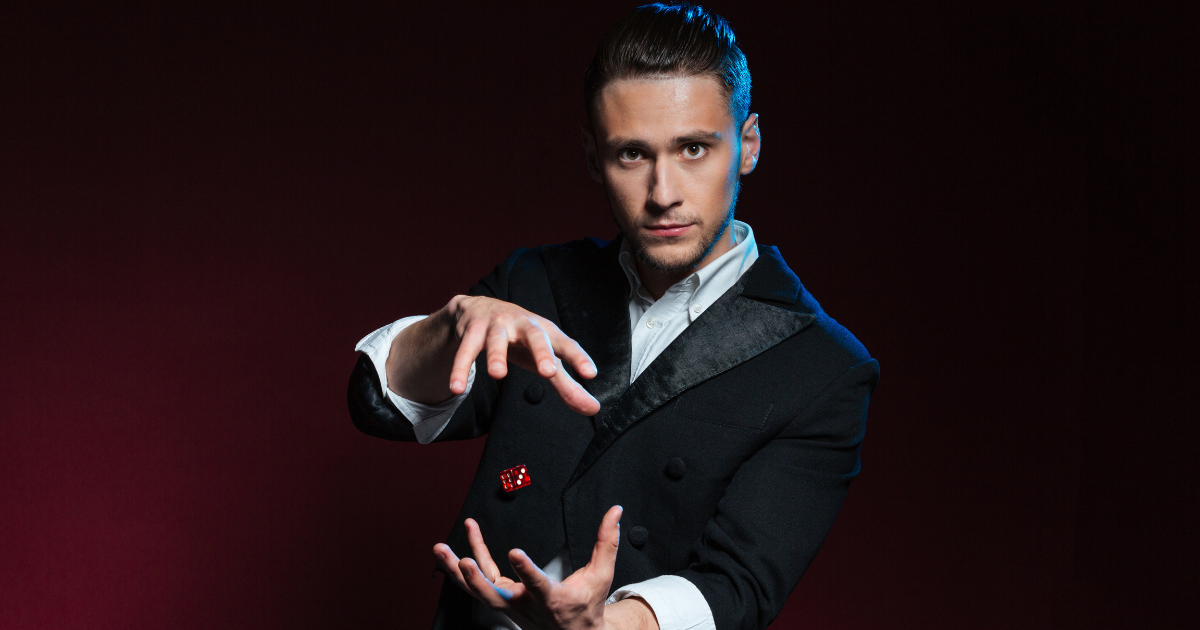 Un homme en costume noir et chemise blanche réalise un tour de magie, semblant faire léviter un dé rouge entre ses mains sur un fond rouge foncé.