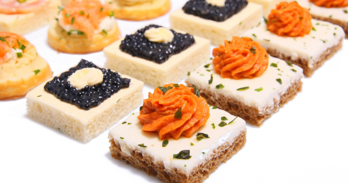 Un assortiment d'élégants canapés de la taille d'une bouchée, comprenant des morceaux de pain carrés garnis de pâte à tartiner crémeuse, de mousse d'orange, d'herbes et de caviar noir, disposés proprement sur un fond blanc.