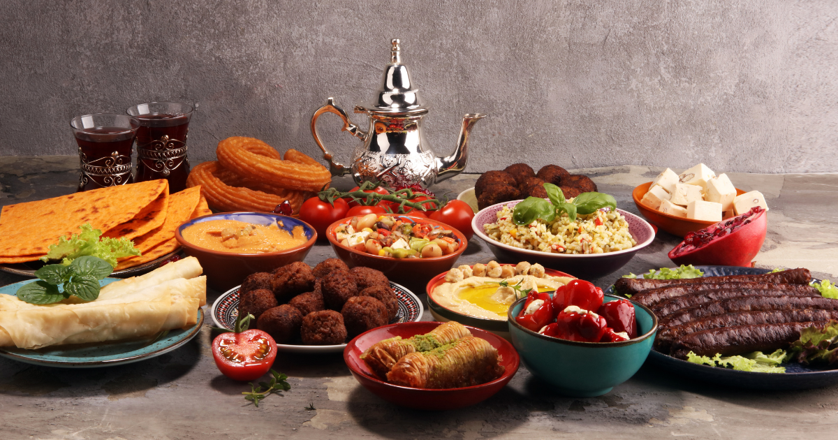 Une table remplie de plats du Moyen-Orient, dont des brochettes, des falafels, du houmous, du taboulé, des légumes frais, du pain, du fromage, du baklava et des churros, avec une théière en argent et des verres de thé à l'arrière-plan.