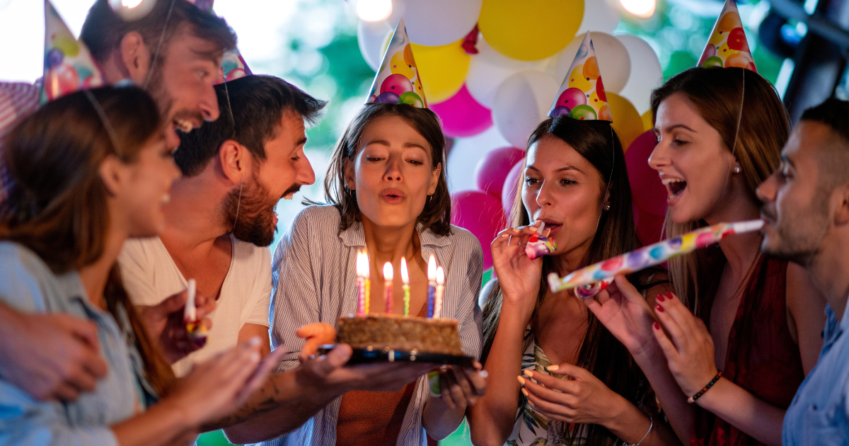 Un groupe d'amis portant des chapeaux de fête célèbre un anniversaire. La personne au centre souffle les bougies sur un gâteau tandis que d'autres sourient, applaudissent et soufflent dans des cornes de fête, avec des ballons colorés en arrière-plan.