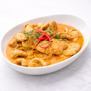 Un plat ovale blanc contient une FILET DE MIGNON DE PORC AU CURRY DOUX dans une sauce crémeuse à l'orange, garnie de fines tranches de piment rouge et d'herbes vertes, le tout posé sur une surface légère.