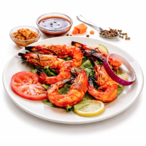 Une assiette de GAMBAS TANDOORI (300 g) est servie sur une salade avec des tranches de tomate, de concombre, de citron et d'oignon rouge, avec des petits bols de sauce, des cornichons et des épices à l'arrière-plan sur une surface blanche.