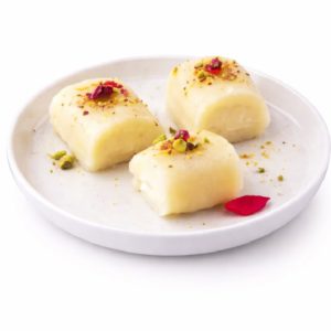 Trois morceaux de HALAWA BIL JIBN, une friandise indienne à base de lait garnie de pistaches et de pétales de rose, sont disposés sur une assiette en céramique blanche sur un fond blanc.