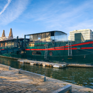 Un grand bateau vert et rouge nommé LE RÊVE FLOTTANT est amarré à une passerelle au bord de la rivière, avec des bâtiments et des parasols en arrière-plan sous un ciel bleu.