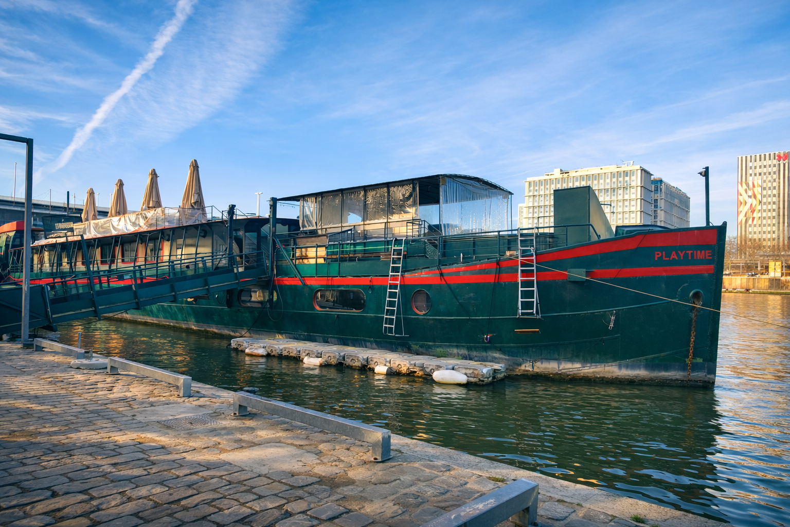 Un grand bateau vert et rouge nommé LE RÊVE FLOTTANT est amarré à une passerelle au bord de la rivière, avec des bâtiments et des parasols en arrière-plan sous un ciel bleu.