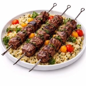 KEFTA DE VIANDE DE BOEUF : Trois brochettes de bœuf grillées sur des brochettes métalliques servies sur du couscous, garnies de persil frais et de tomates cerises. Commande minimum : 5 assiettes de 300g par personne.