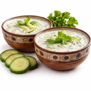 Deux bols LABAN (250g) remplis de soupe de concombre à base de yaourt, garnis de feuilles de menthe et de tranches de concombre. Du persil frais et des tranches de concombre supplémentaires sont disposés à côté des bols sur un fond blanc.