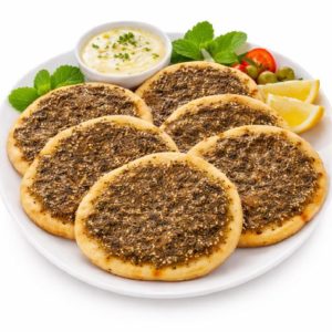 Une assiette blanche contenant six MANAKISH ZAATAR (80 g/pièce) est servie avec des quartiers de citron, des olives, des tomates, des feuilles de menthe et un petit bol de sauce crémeuse aux herbes.
