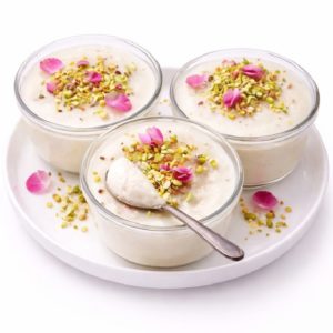 Trois coupes en verre de MOUHALLABIEH FLAN À LA FLEUR D'ORANGER, garnies de pistaches hachées et de pétales de roses, sont disposées sur une assiette blanche. Une cuillère est posée dans l'un des bols.