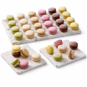 Le PLATEAU DE 72 MACARONS EN ASSORTIMENT présente un assortiment de macarons colorés, roses, jaunes, bruns, crème et verts, disposés proprement sur trois dalles de pierre, en rangées et en petits groupes.