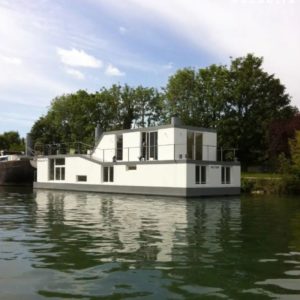 LA PETITE PÉNICHE DE SÈVRES (92310), une péniche blanche moderne avec de grandes fenêtres, flotte sur des eaux calmes près d'un rivage luxuriant bordé d'arbres, sous un ciel partiellement nuageux.