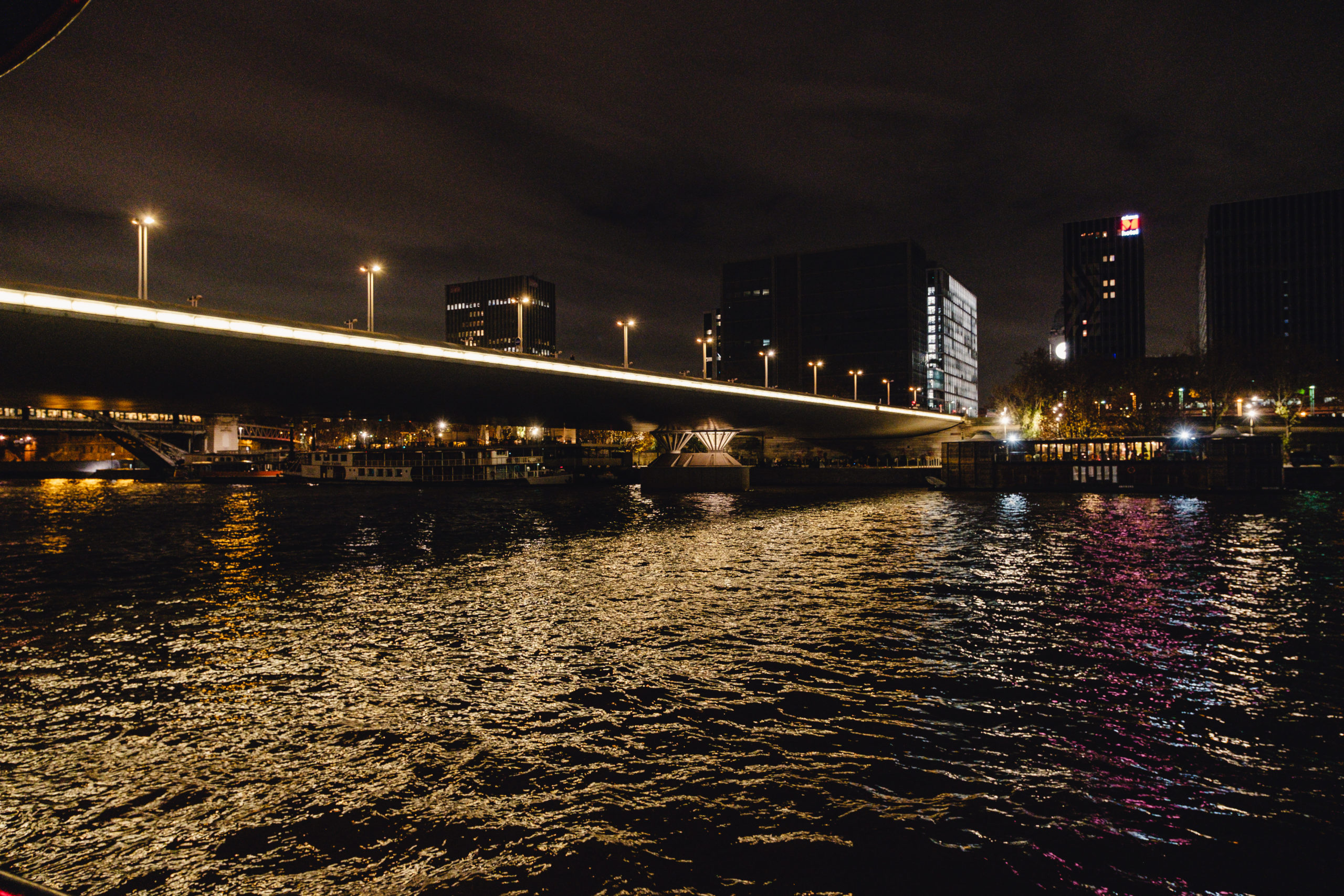 LE RÊVE FLOTTANT capture un pont urbain éclairé de nuit au-dessus d'une rivière, les lampadaires se reflétant sur l'eau. Des bâtiments modernes illuminés se dressent à l'arrière-plan sous un ciel nuageux.