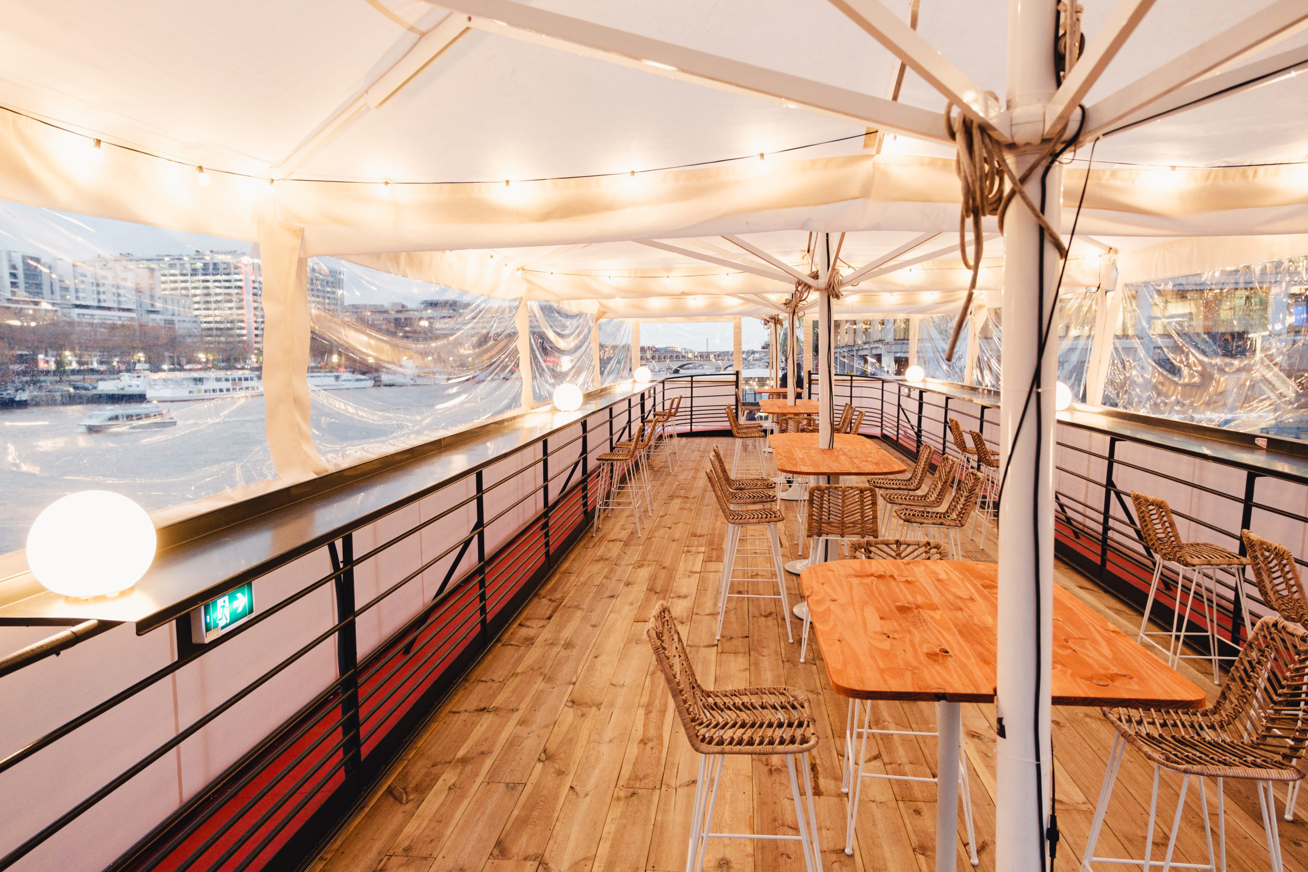 LE RÊVE FLOTTANT propose une salle à manger couverte sur un bateau avec des tables en bois, des chaises en osier, des guirlandes lumineuses et des vues de la ville sur le fleuve à travers des rideaux en plastique transparent.