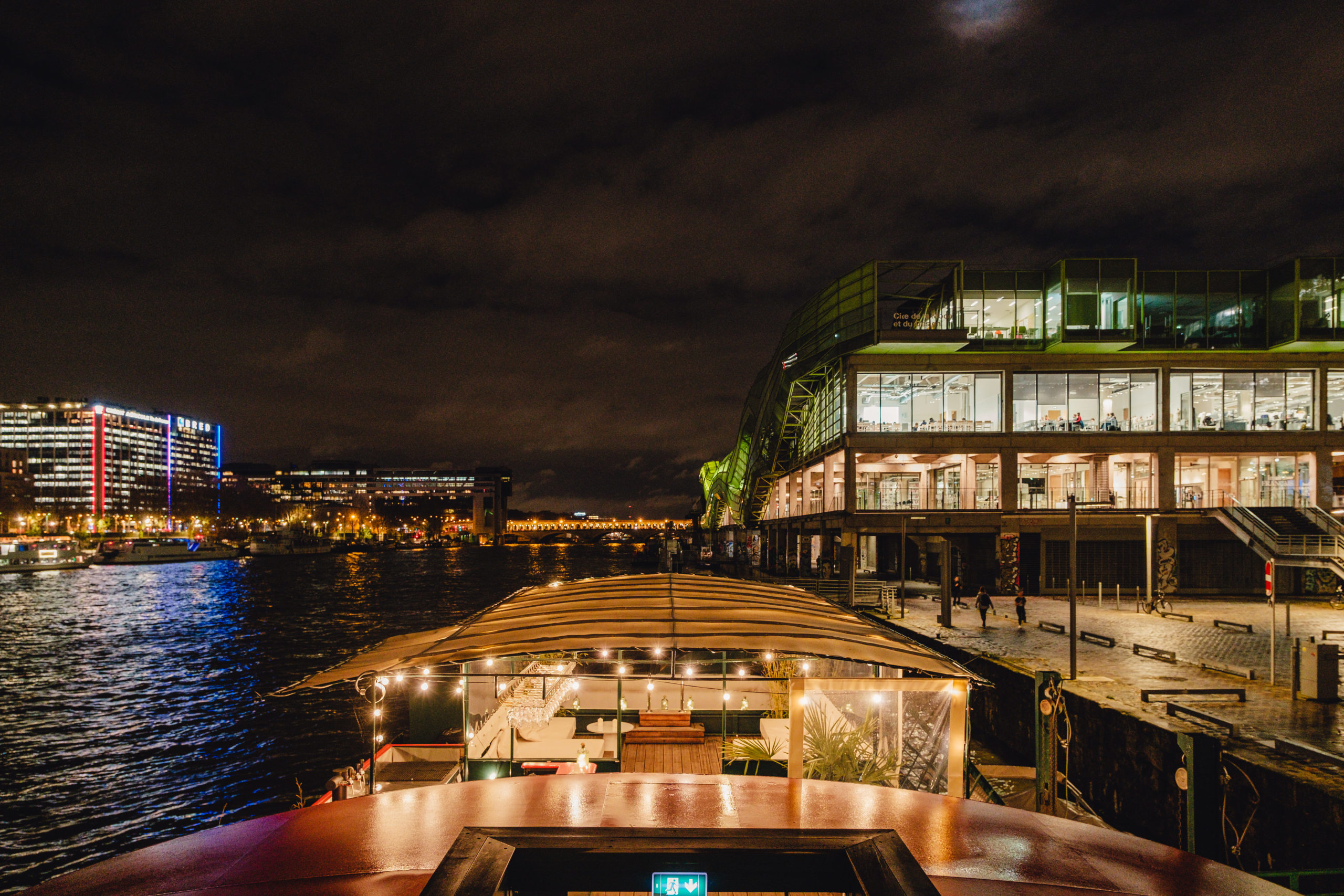 LE RÊVE FLOTTANT : une vue nocturne de la rivière avec les lumières de la ville se reflétant sur l'eau, un bateau décoré amarré à l'avant, et un bâtiment moderne très éclairé avec de grandes fenêtres à droite, sous un ciel nuageux.