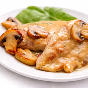 SUPRÊME DE VOLAILLES AUX CHAMPIGNONS : Suprême de volaille aux champignons et sauce crémeuse, servi sur une assiette blanche avec des feuilles d'épinards frais en arrière-plan.