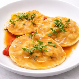 Trois GROS RAVIOLIS AU HOMARD à la sauce orange légère sont garnis d'herbes fraîches et servis sur une assiette blanche, avec des morceaux de tomates visibles en dessous.