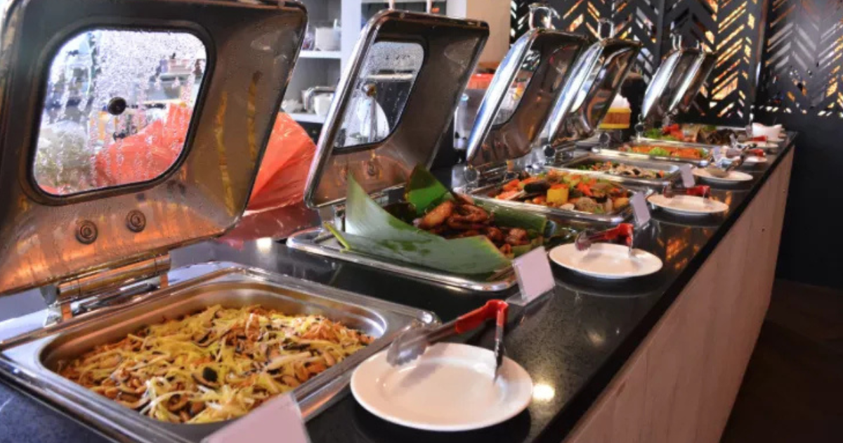 Un buffet composé de divers plats chauds dans des chafing dishes en acier inoxydable, chacun avec un ustensile de service et une assiette blanche devant, posés sur un comptoir noir dans un restaurant bien éclairé.
