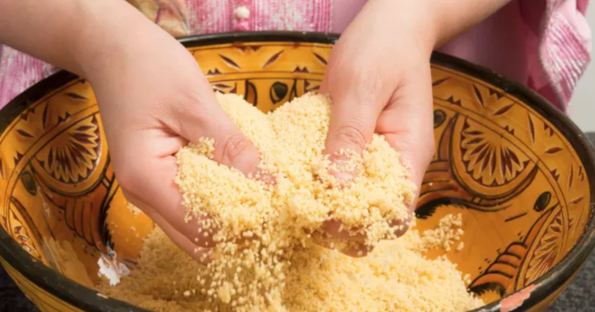 Deux mains mélangent des grains de couscous jaune dans un bol décoratif à motifs orange et marron.