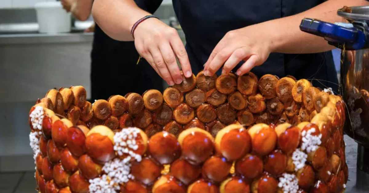 Une personne assemble un grand croquembouche en empilant soigneusement des choux à la crème glacés au caramel selon un schéma circulaire dans une cuisine.