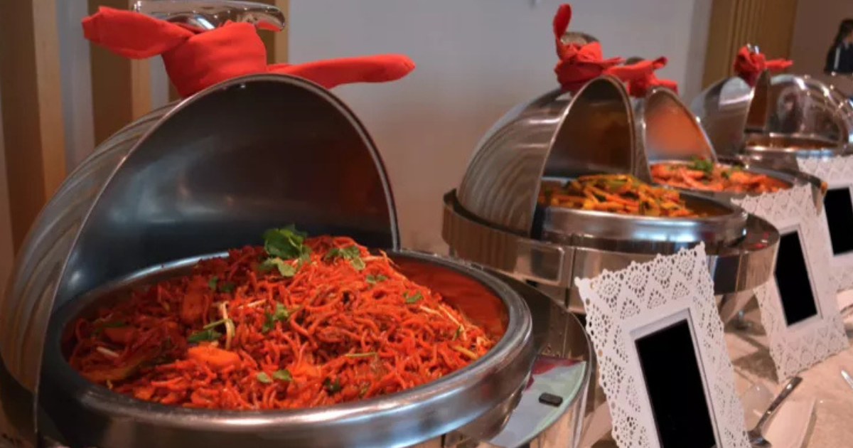 Buffet composé de plusieurs chafing dishes couverts, remplis de nouilles colorées et garnis d'herbes ; chaque plat comporte une serviette rouge sur le couvercle et une étiquette vierge sur le devant.
