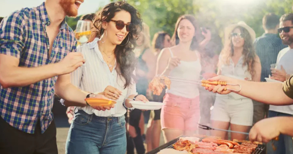 Un groupe de personnes se réunit en plein air autour d'une grille de barbecue, souriant et servant de la nourriture. Une personne tient une assiette, une autre sert de la viande, tandis que d'autres discutent en arrière-plan, profitant d'une journée ensoleillée.