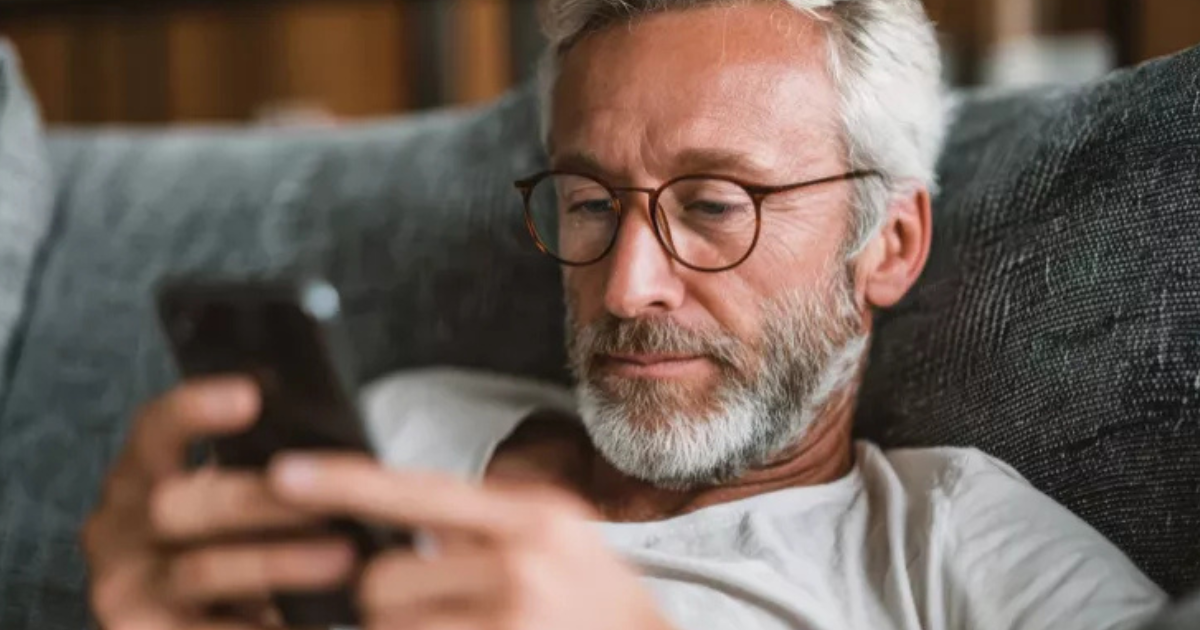 Un homme d'âge moyen avec des cheveux gris, une barbe et des lunettes est allongé sur un canapé, regardant attentivement un smartphone qu'il tient à deux mains.