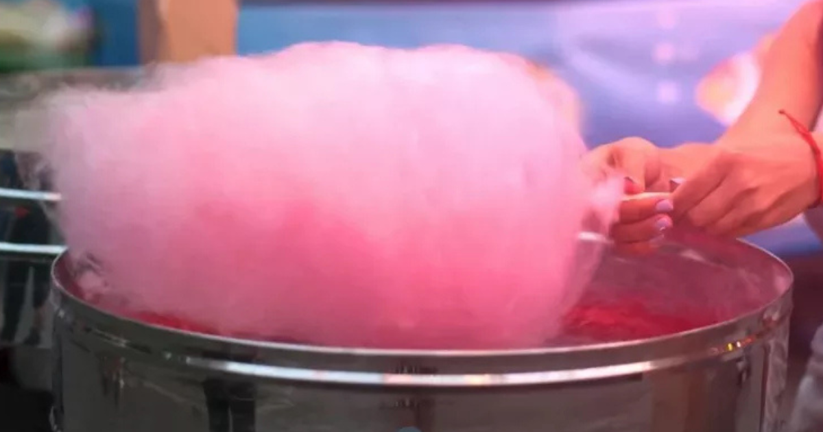 Une personne fait tourner un gros nuage rose et floconneux de barbe à papa sur un bâton au-dessus d'une machine à barbe à papa en métal. L'arrière-plan est flou et coloré.