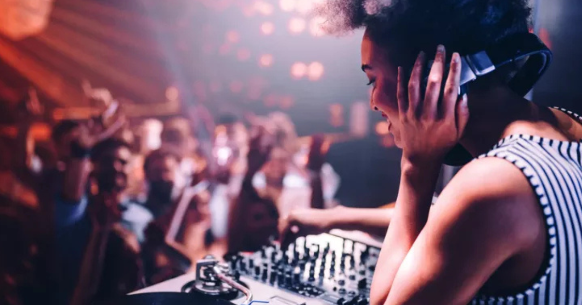 Un DJ muni d'un casque mixe de la musique sur une table tournante, tandis qu'une foule animée danse et lève les mains en arrière-plan sous des lumières colorées.