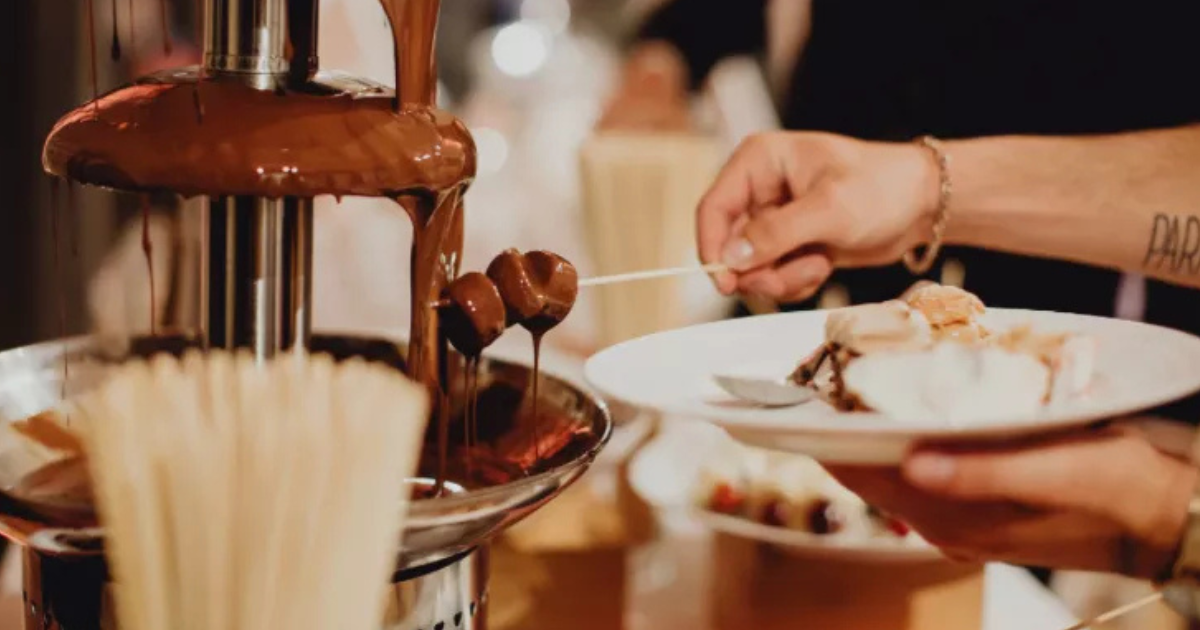 Une personne trempe des brochettes dans une fontaine de chocolat qui coule, tout en tenant une assiette de desserts dans l'autre main.