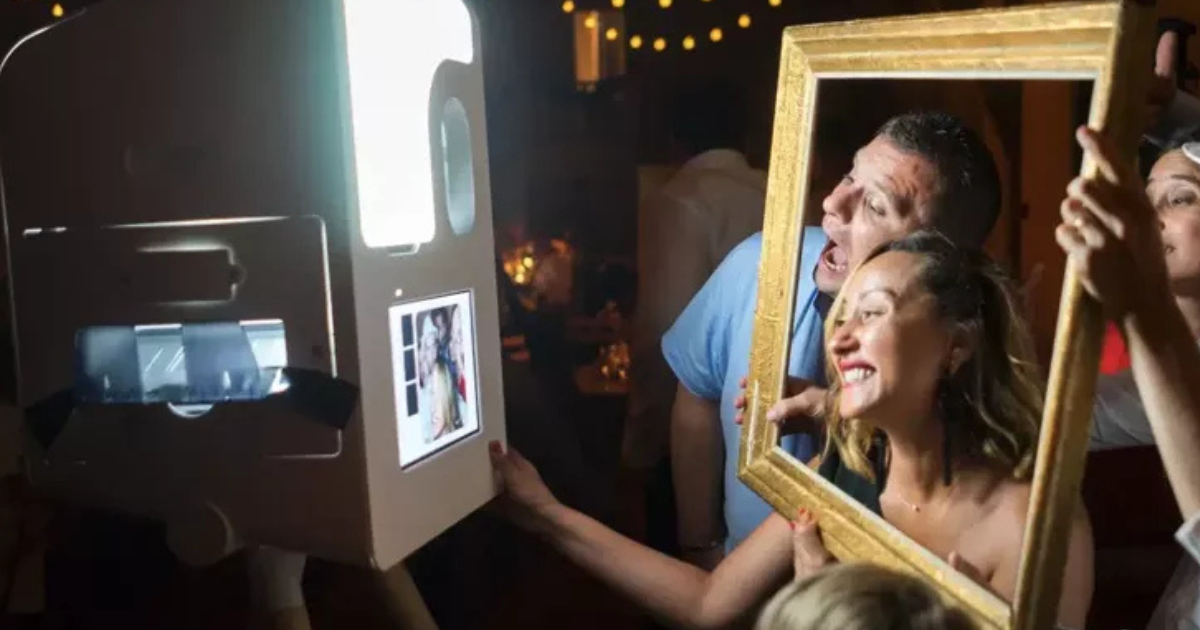 Deux personnes sourient et posent derrière un cadre doré devant un photomaton lors d'un événement animé en intérieur, avec des guirlandes lumineuses et d'autres invités en arrière-plan.