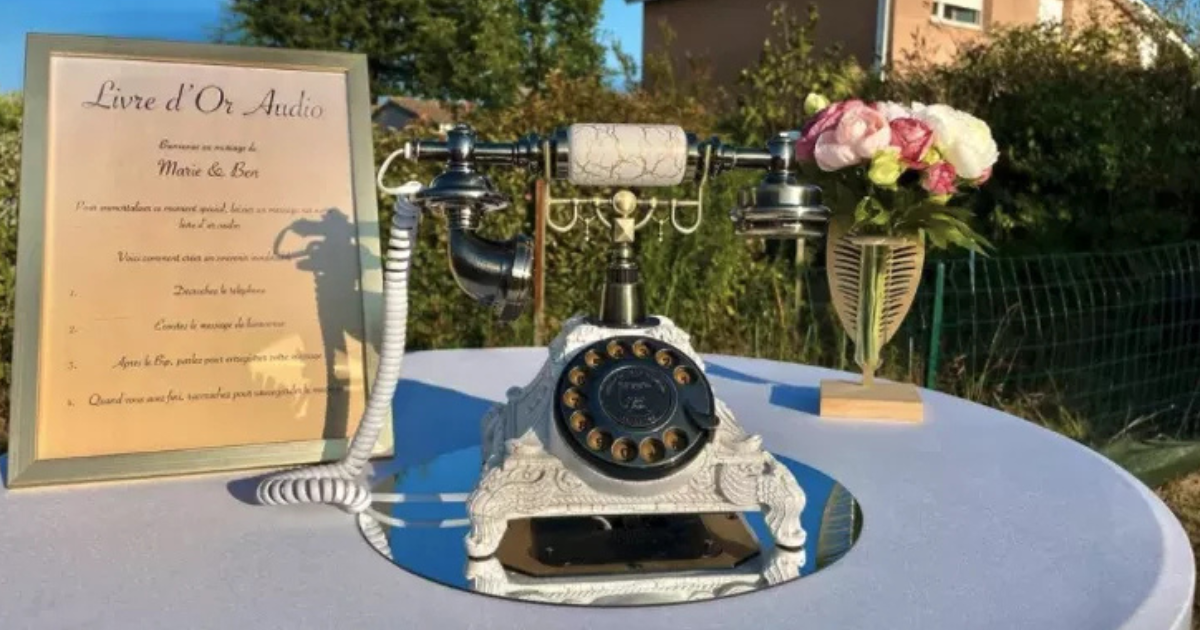 Un téléphone rotatif vintage est posé sur un plateau en miroir à côté d'un panneau encadré et d'un vase de fleurs sur une table ronde drapée d'une nappe blanche, placée en extérieur avec de la verdure et une maison en arrière-plan.