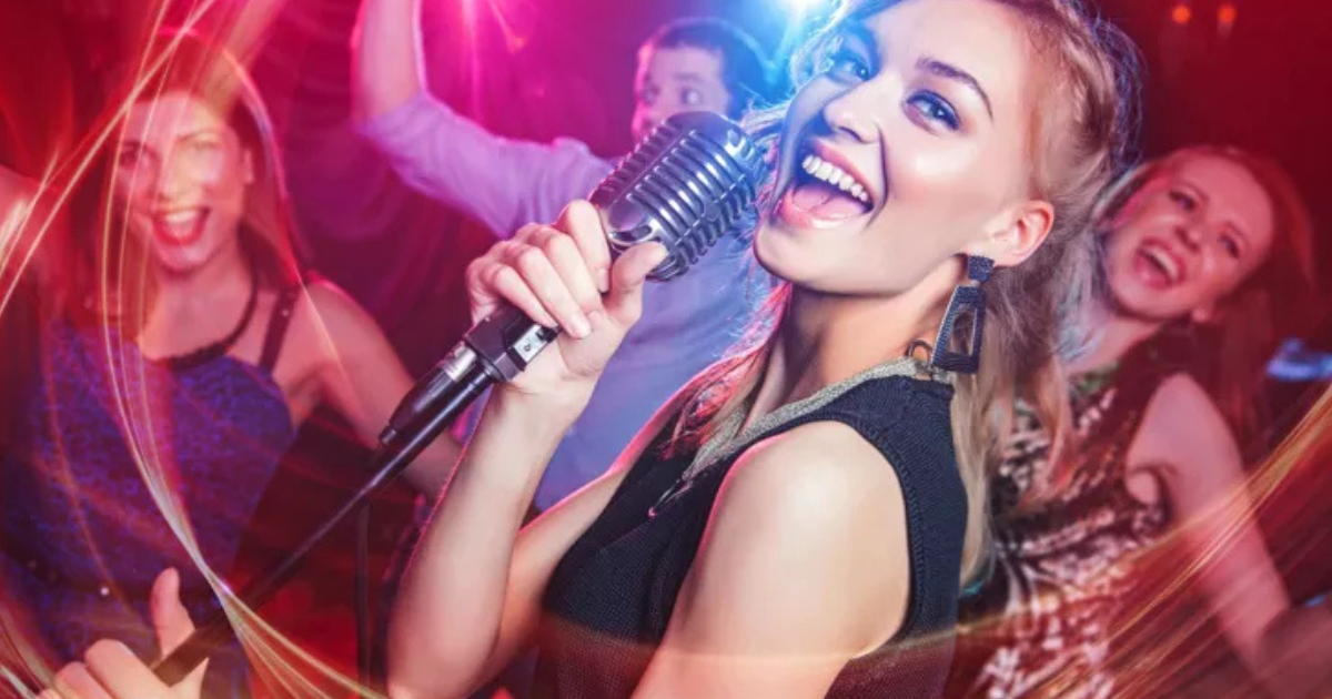 Une femme chante dans un micro sur scène, souriant énergiquement, avec des amis qui dansent et l'acclament derrière elle, des lumières colorées ajoutant une atmosphère de fête.