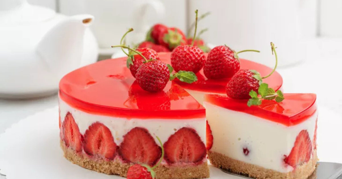 Un cheesecake rond aux fraises avec une couche de gelée rouge brillante sur le dessus, décoré de fraises fraîches et de menthe. Une tranche a été coupée, révélant des fraises entières à l'intérieur de la garniture crémeuse et une base de biscuit friable.