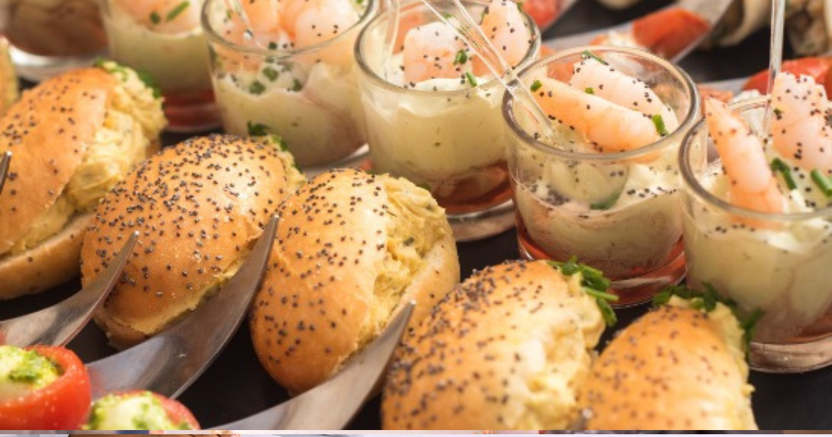 Un assortiment d'amuse-gueules, dont des mini-sandwichs au pavot, des cocktails de crevettes servis dans de petits verres et des tomates farcies, disposés sur un plateau.