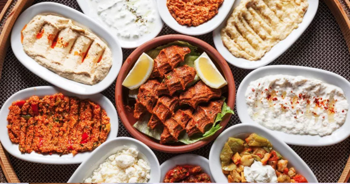 Une variété de plats mezes libanais disposés dans de petites assiettes ovales autour d'une assiette centrale de viande tranchée garnie de quartiers de citron, avec des trempettes, des pâtes à tartiner et des salades sur un plateau rond.