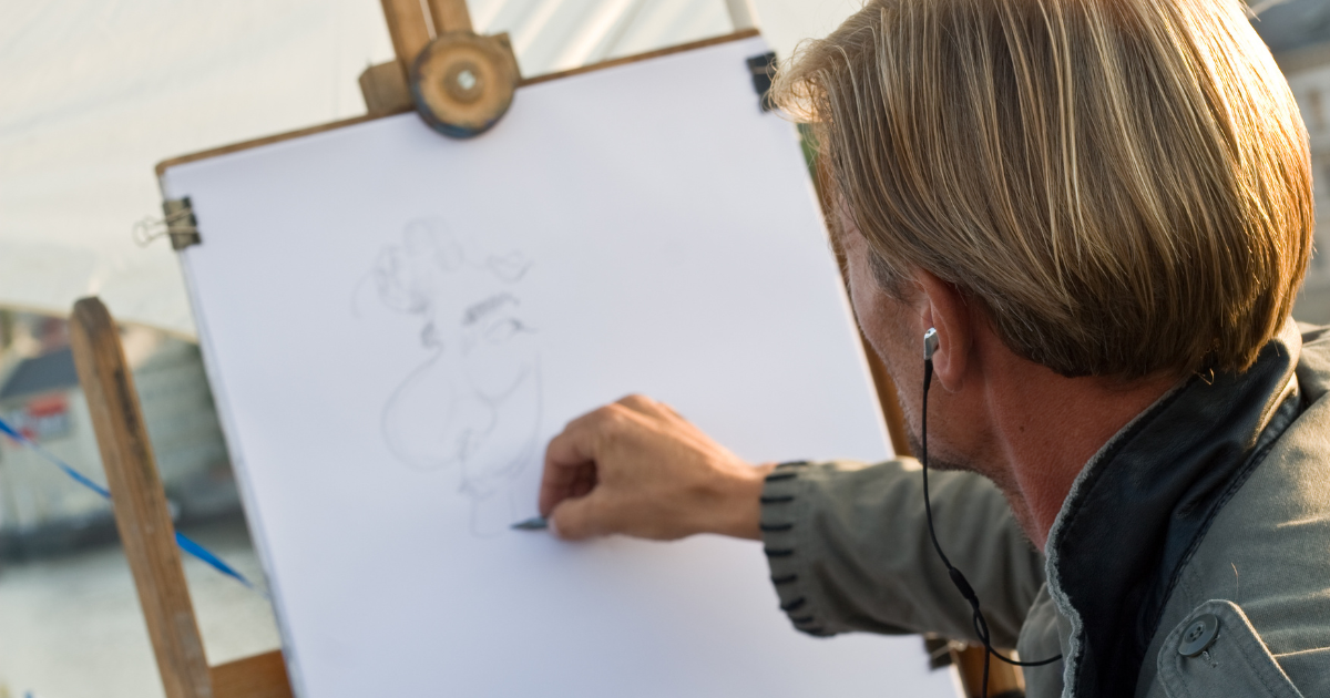 Une personne aux cheveux blonds esquisse une caricature sur une grande feuille blanche, à l'aide d'un crayon et d'écouteurs. Le chevalet de dessin est installé en plein air.