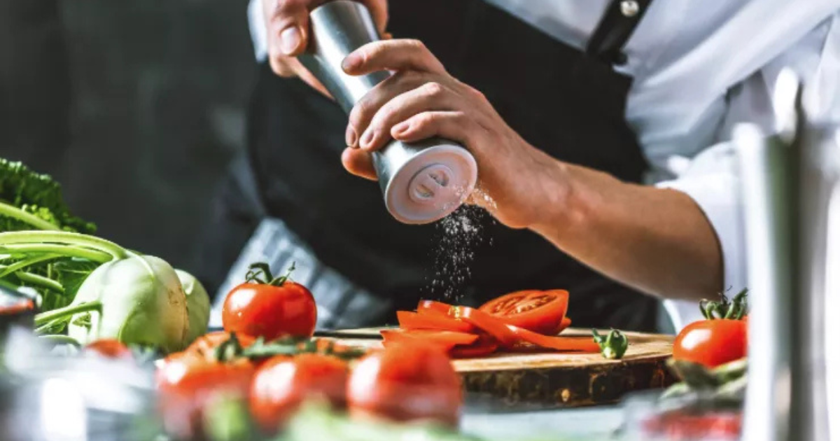 Une personne vêtue d'un tablier sombre saupoudre le sel d'une salière métallique sur des tranches de tomates posées sur une planche à découper en bois, entourée de légumes frais.