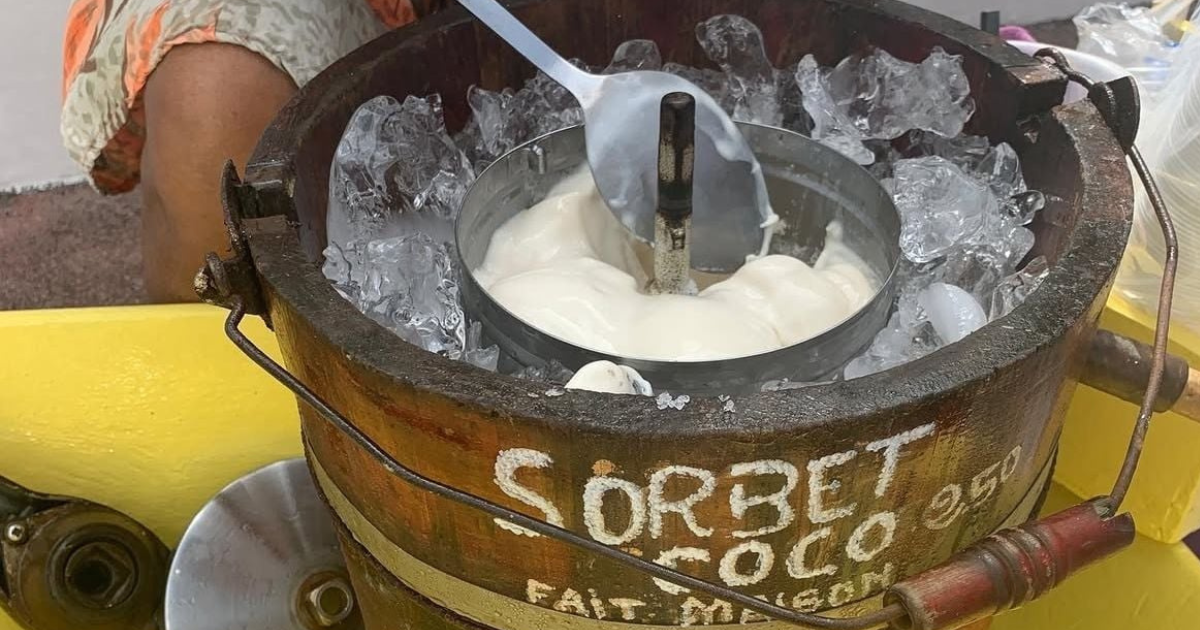 Gros plan sur une baratte à glace en bois remplie de sorbet à la noix de coco entouré de glace. Une louche en métal repose à l'intérieur et les mots SORBET COCO sont peints sur la baratte. Les mains et les vêtements colorés d'une personne sont partiellement visibles.