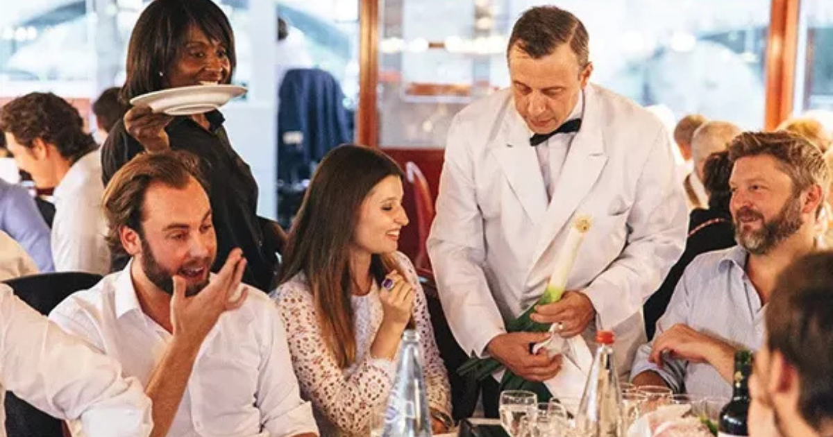 Un serveur en veste blanche sert une table de convives souriants dans un restaurant très fréquenté, tandis qu'une serveuse en noir se tient à proximité en tenant des assiettes. La table est garnie de verres, de bouteilles et de couverts.