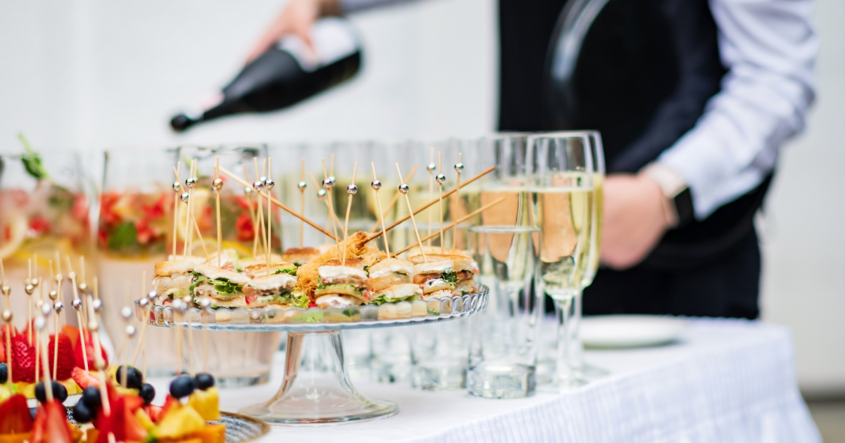 Lors d'un événement festif, une personne verse du champagne dans des coupes sur une table blanche garnie d'amuse-gueule, notamment de sandwichs en brochettes et de fruits.