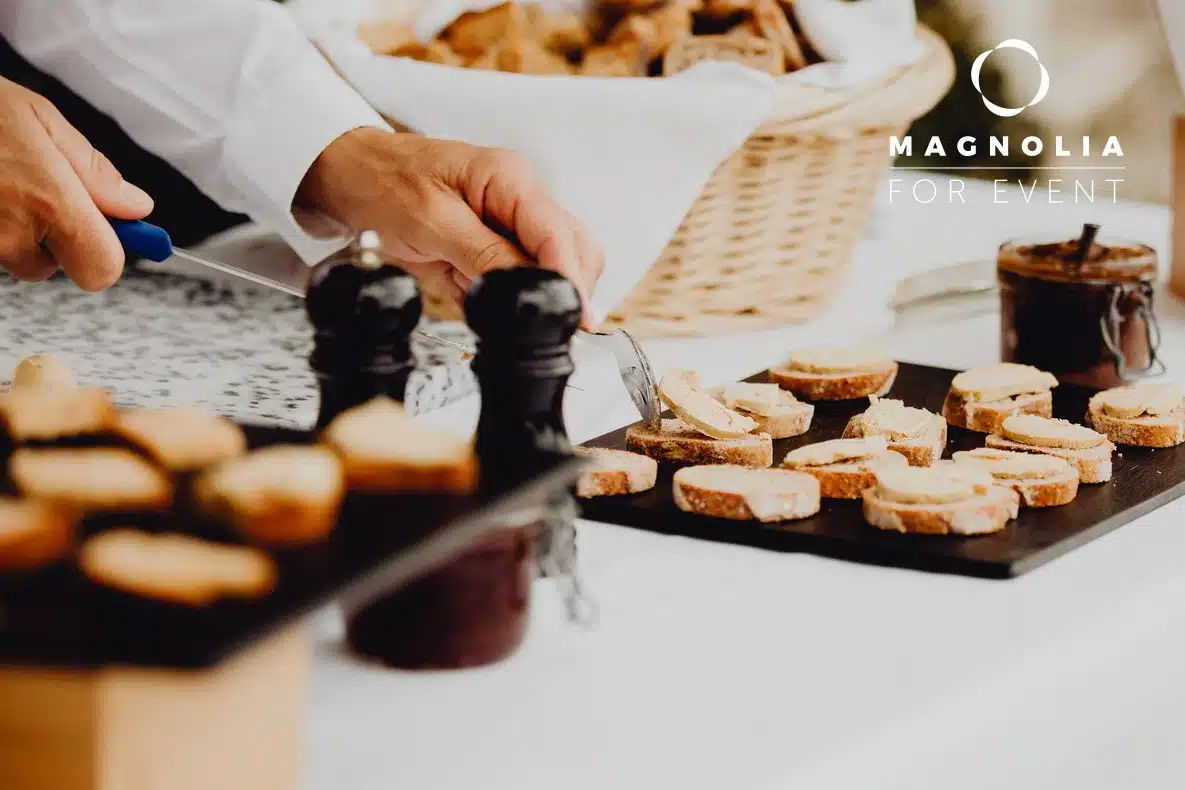 Une personne dispose des tranches de pain avec de la pâte à tartiner sur un plateau, avec des bocaux et des moulins à poivre à proximité. À l'arrière-plan, on aperçoit une corbeille contenant d'autres pains. L'image comprend le texte "Magnolia For Event".