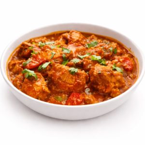 Un bol blanc d'AGNEAU TIKKA (300 g) présente un riche curry rouge avec de tendres morceaux de viande, des tomates et une garniture de coriandre fraîche.