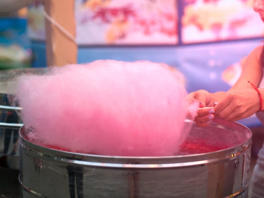 Une personne fabriquant de la barbe à papa rose à une machine, avec une main tenant un bâton et faisant tourbillonner la barbe à papa duveteuse, sur un fond coloré flou.