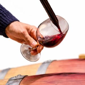 Une main incline un verre de vin tandis que le vin rouge est versé, avec des tonneaux en bois à proximité - faites-en l'expérience avec l'ANIMATION OENOLOGIE.