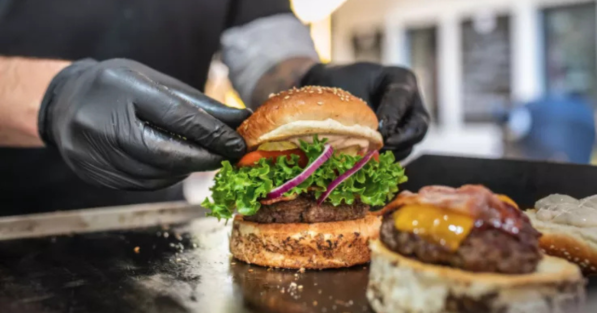 Une personne portant des gants noirs prépare un hamburger avec de la laitue, de la tomate et de l'oignon rouge, en plaçant le pain sur le dessus. Un autre hamburger avec du fromage fondu et du bacon est posé sur le côté, tous deux sur un gril ou un poste de préparation.