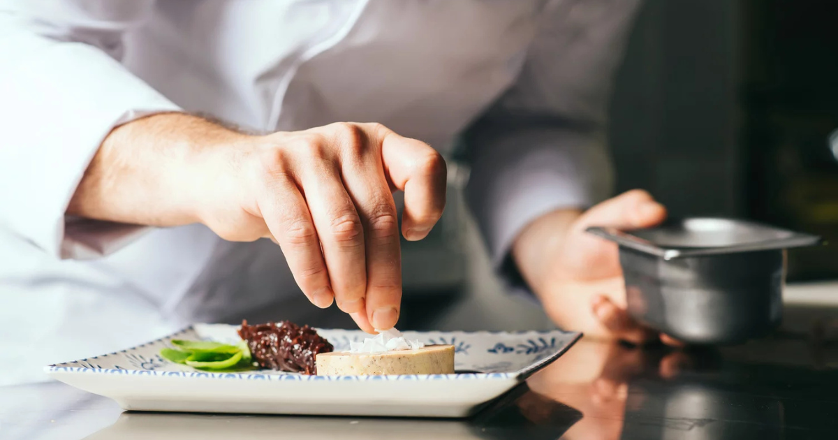 Un chef en blouse blanche saupoudre délicatement une garniture sur un plat qui contient également une sauce et une garniture verte sur une assiette à motifs.