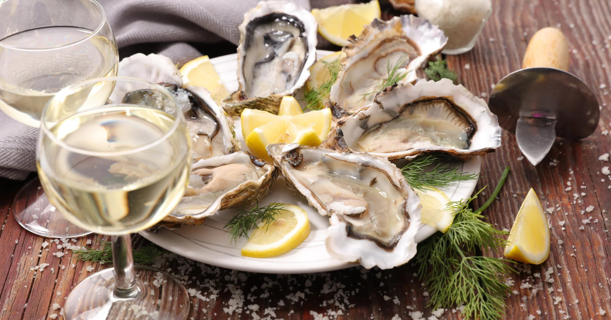 Une assiette d'huîtres fraîches sur la demi-coquille, garnies de quartiers de citron et d'aneth, entourées de deux verres de vin blanc sur une table en bois rustique saupoudrée de gros sel.