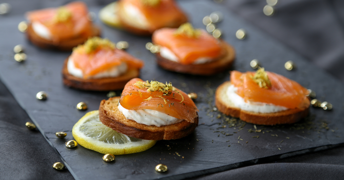Des tranches de pain grillé garnies de fromage frais, de saumon fumé et d'herbes sont disposées sur un plateau de service en ardoise avec des tranches de citron et des perles décoratives dorées.