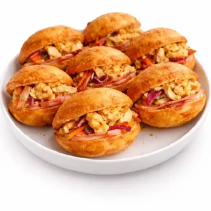 Une assiette blanche contient sept MINI BOKIT GARNI POULET (80 g chacun), des petits pains ronds farcis de poulet, d'œufs brouillés, de carottes râpées et d'oignons rouges, soigneusement disposés sur un fond blanc. Commande minimum : 7 pièces.